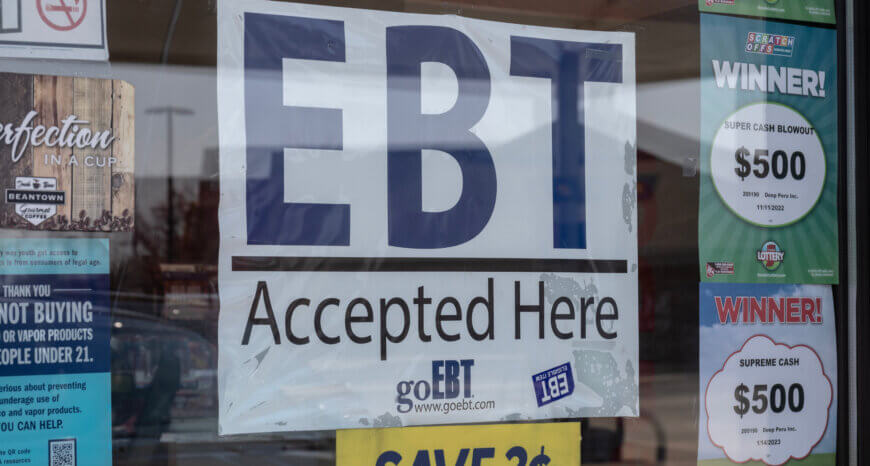 A storefront window displaying a large EBT Accepted here sign in the center of smaller ads